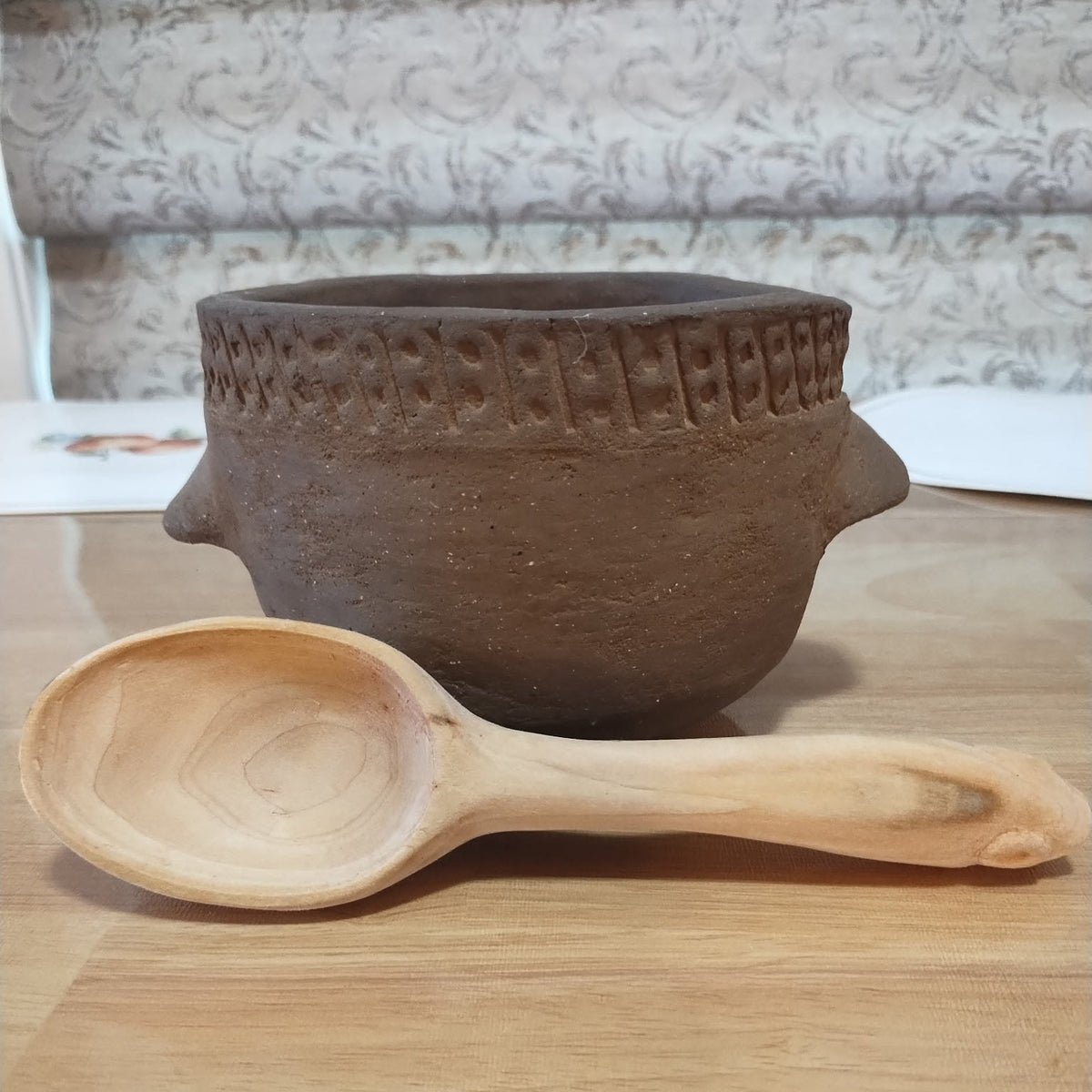 A hand-carved light wood spoon resting in front of a dark brown earthenware pot with a decorative rim, set on a light wooden tabletop.