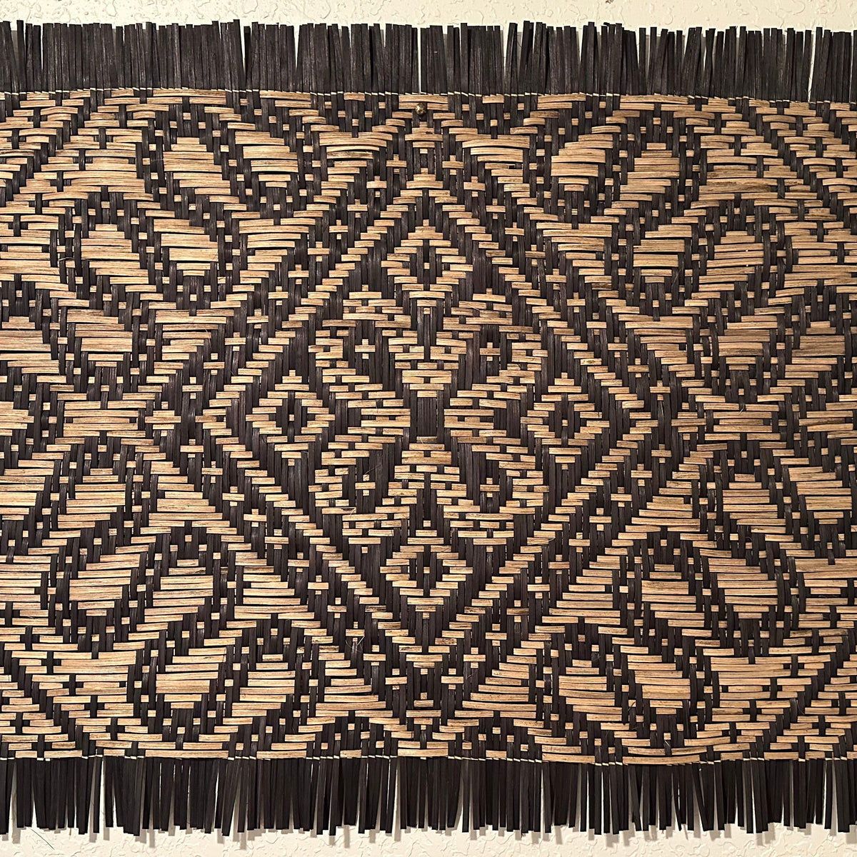 A rectangular woven wall hanging or mat with a geometric black and tan pattern and dark fringe on all sides, mounted against a light beige wall.