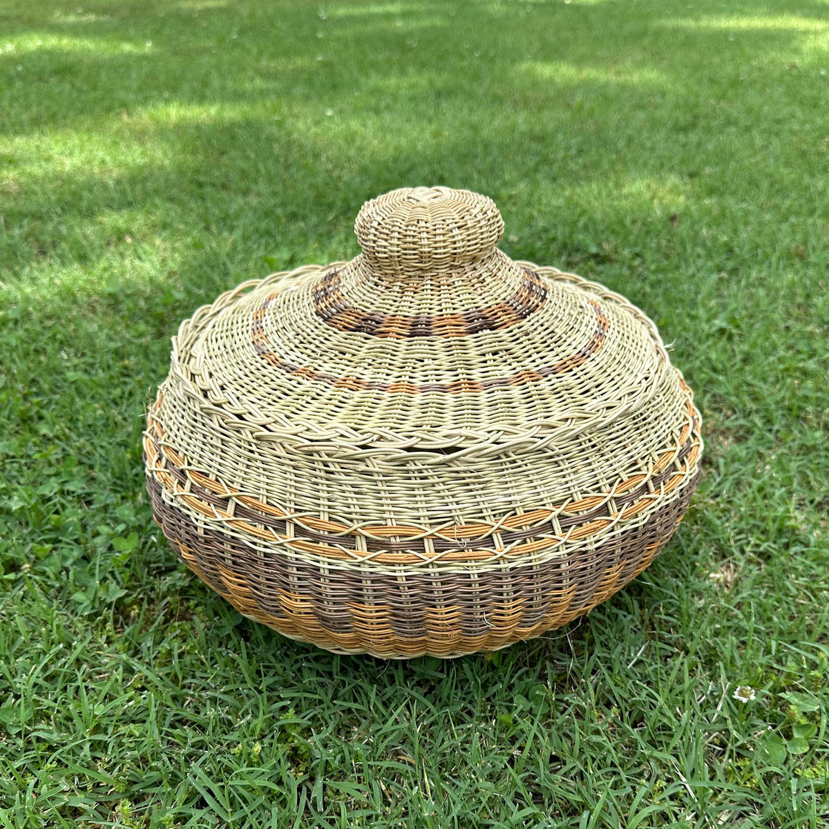 A round, hand-woven basket with a matching lid sits on a green grass lawn.
