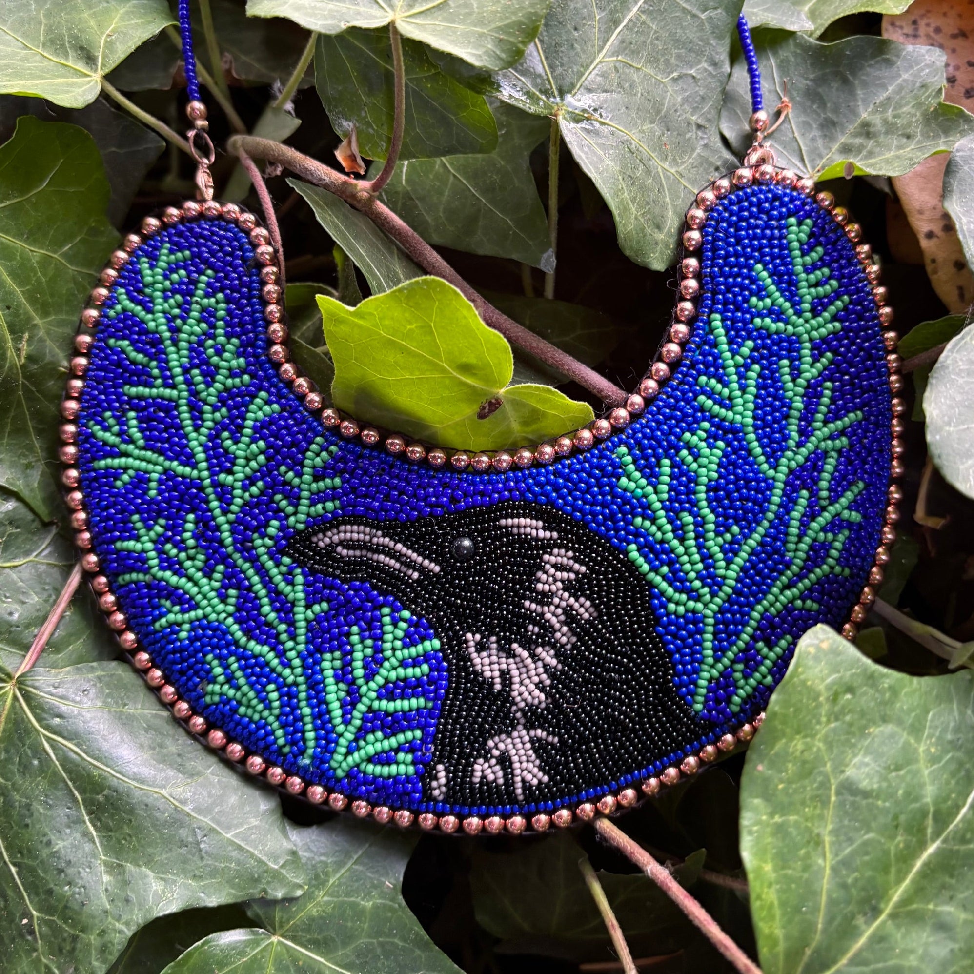 A handmade beaded crescent-shaped necklace featuring a raven design and green leaf patterns on a vibrant blue background, styled among green ivy leaves.