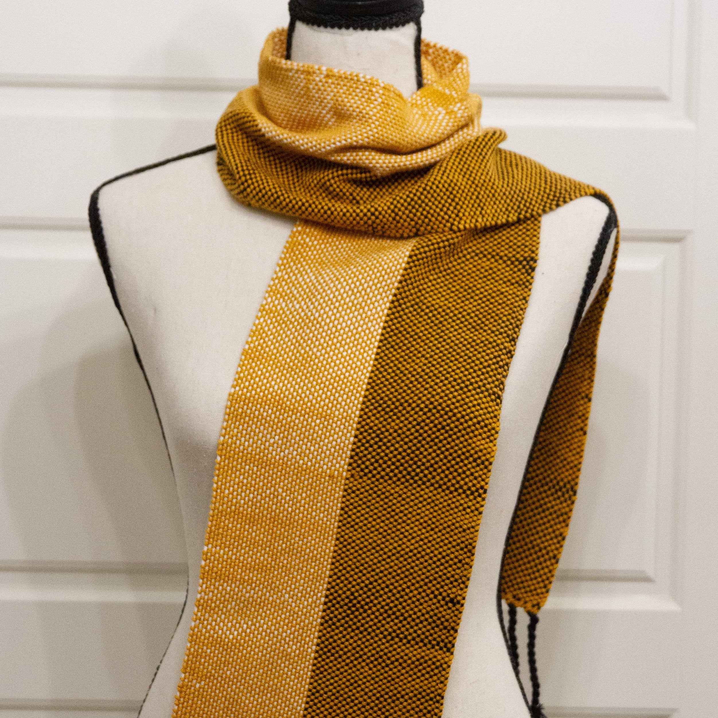 A mustard yellow and dark brown woven scarf with white and black fringe tassels, displayed on a cream-colored dress form mannequin against a white paneled door background.